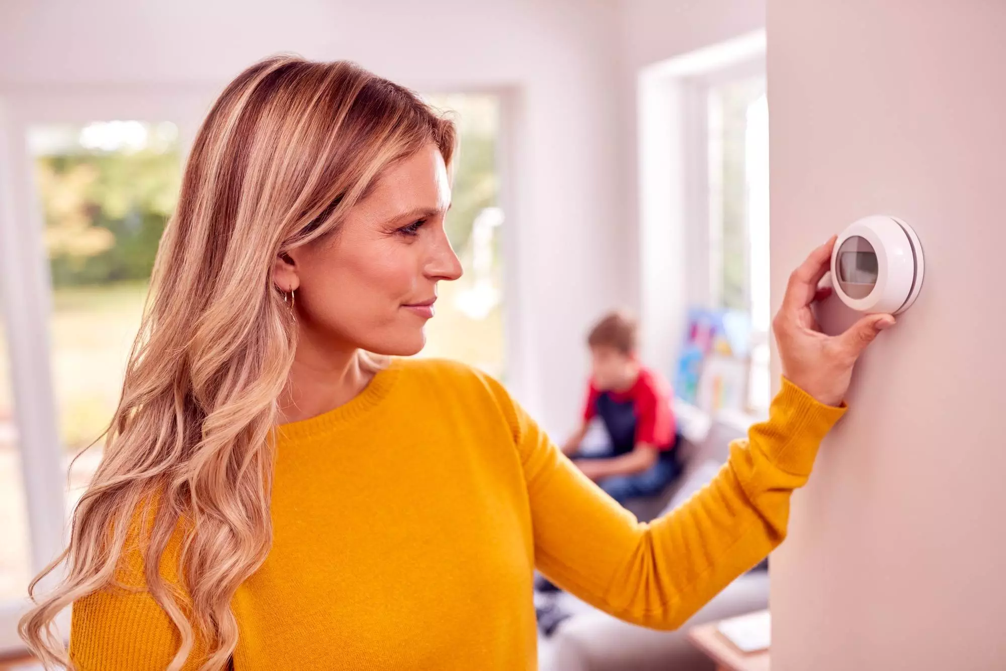 A woman in a yellow sweater adjusts a digital thermostat on the wall, ensuring ideal comfort with Horizon Air's expert AC Installation, while a child sits in the background of a bright living room.